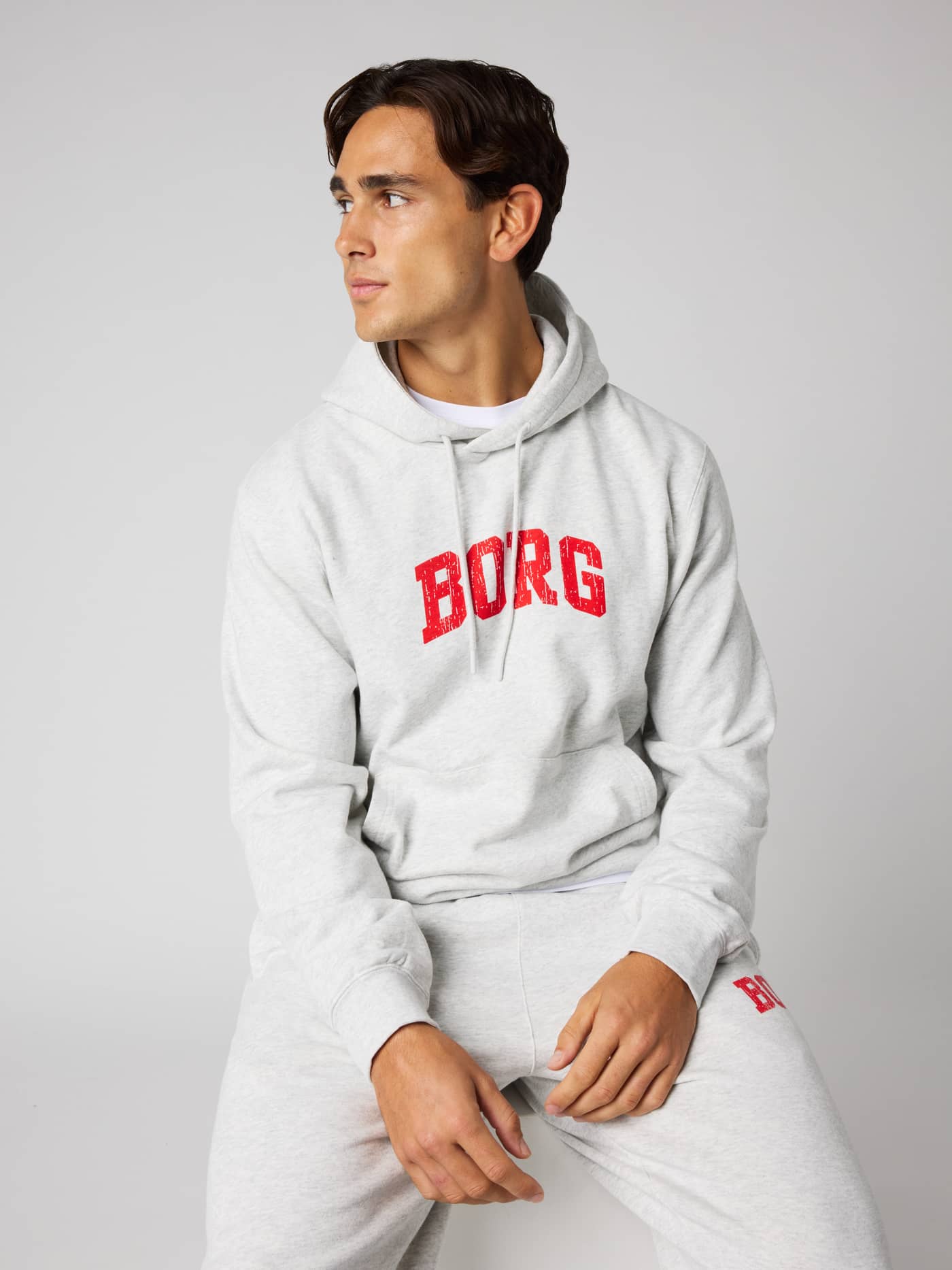 A man sitting in the Borg Classic Print Hoodie in Ultra Light Grey Melange (Grey) with a red logo for tennis and training. - Image 4
