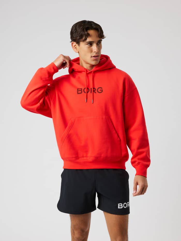 Vue de face d'un homme portant le Borg Oversized Hoodie en Flame Scarlet (Rouge) avec logo noir pour l'entraînement.
