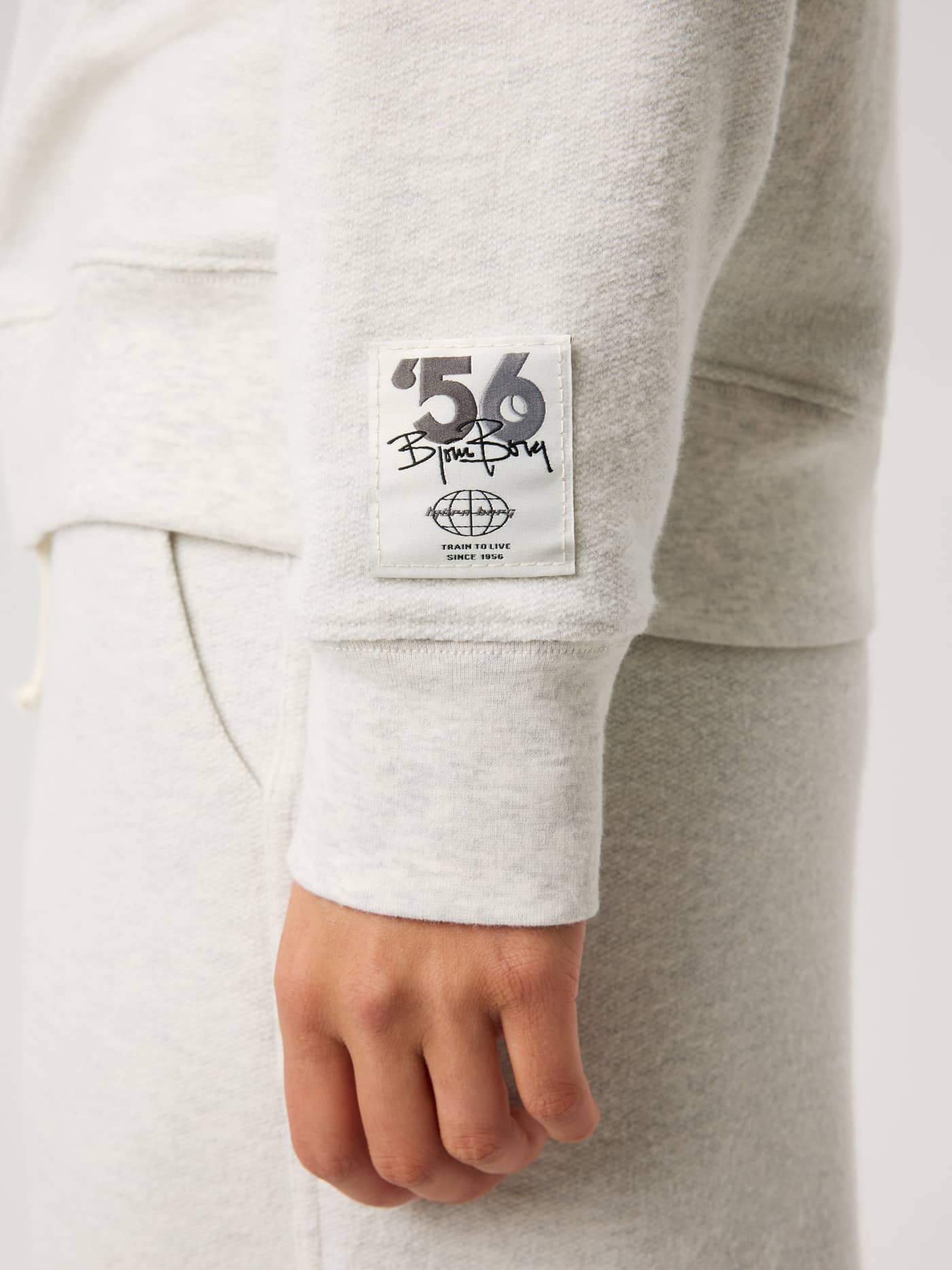 Close-up of the Studio French Terry Oversized Sweatshirt in Tofu Melange (Beige) featuring a black logo patch on the sleeve. - Image 5