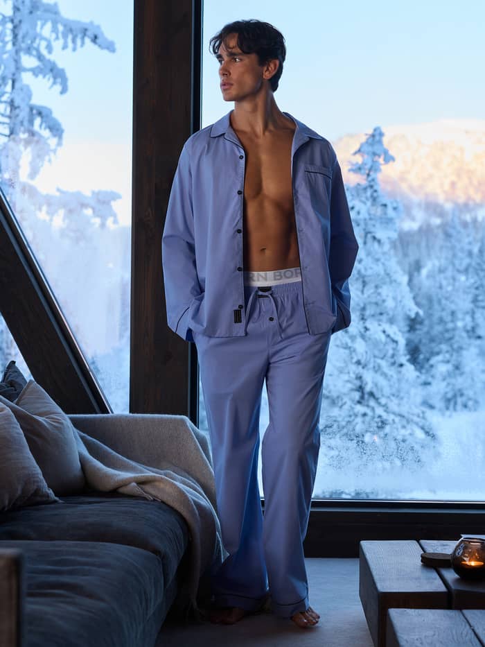 En mand iført Core Premium Pyjama Pants i Lavender Lustre (blå) indendørs nær et snedækket vindue.