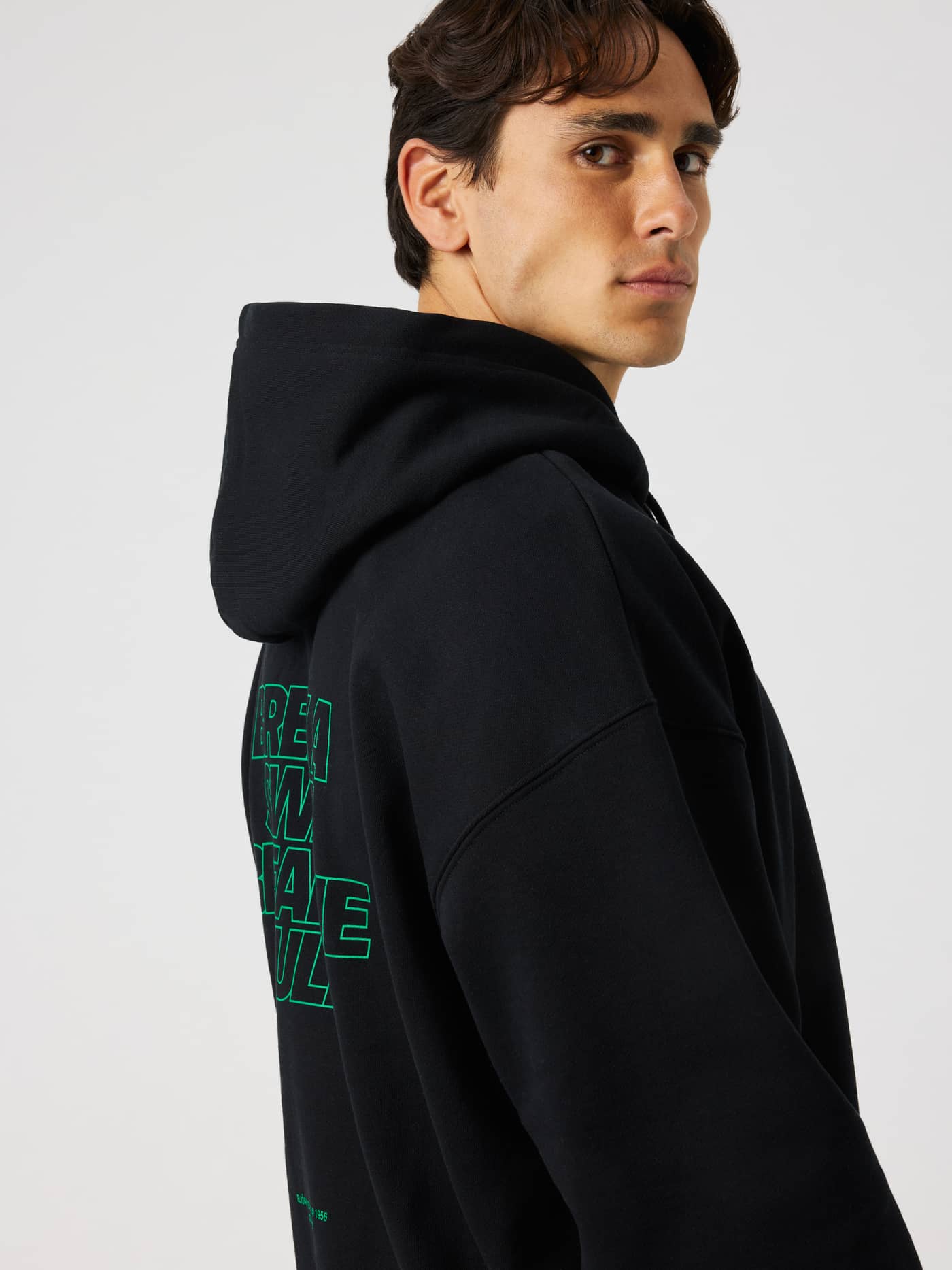 Side profile of a model wearing the Borg Oversized Hoodie in Black Beauty (Black) featuring green text on the back. - Image 1