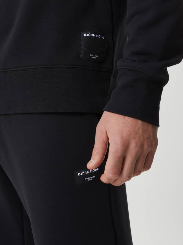 Detailed close-up of the soft cotton Centre Crew in Black Beauty (Black) featuring a white woven logo label on the hip. - Image 5