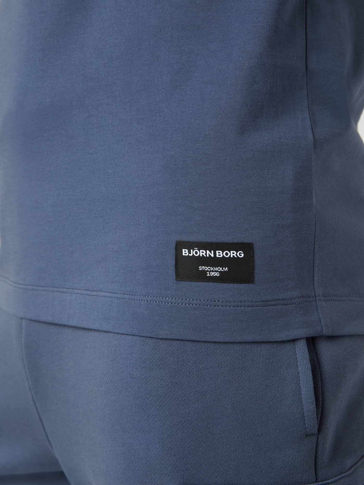 Detailed close-up of the men's Centre T-Shirt in Nightshadow Blue (Blue) with a black woven logo label at the hem. - Image 4
