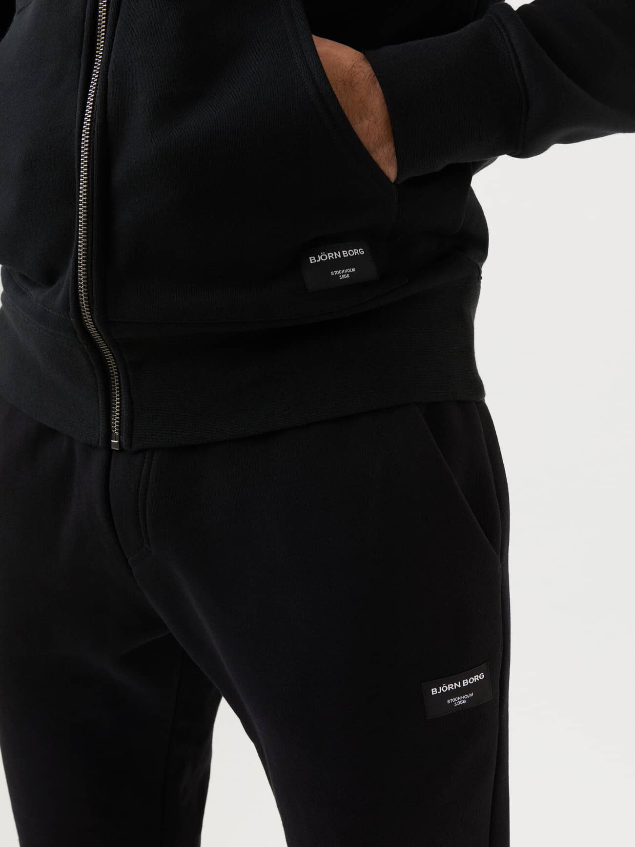 Close-up of the Centre Zip Hood in Black Beauty (Black), showcasing functional pockets and white woven logo labels. - Image 3