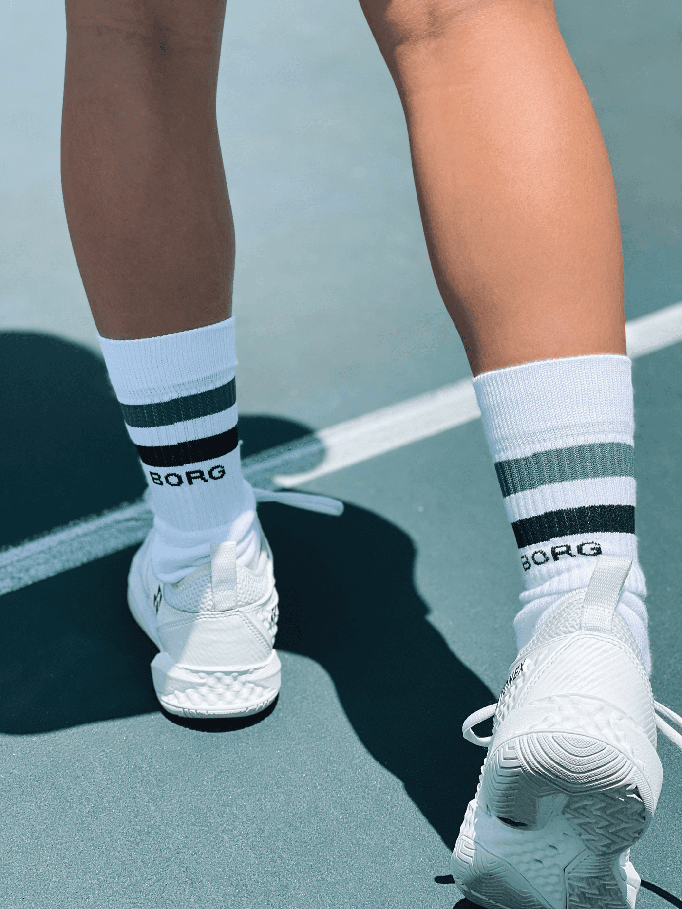 Back view of a model wearing white Björn Borg Core Crew Socks with green and navy stripes and a navy logo on a tennis court. - Image 2