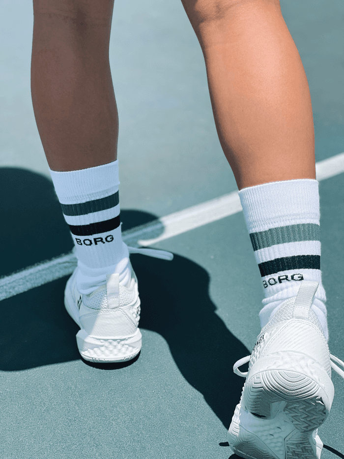 Back view of a model wearing white Björn Borg Core Crew Socks with green and navy stripes and a navy logo on a tennis court. - Image 2