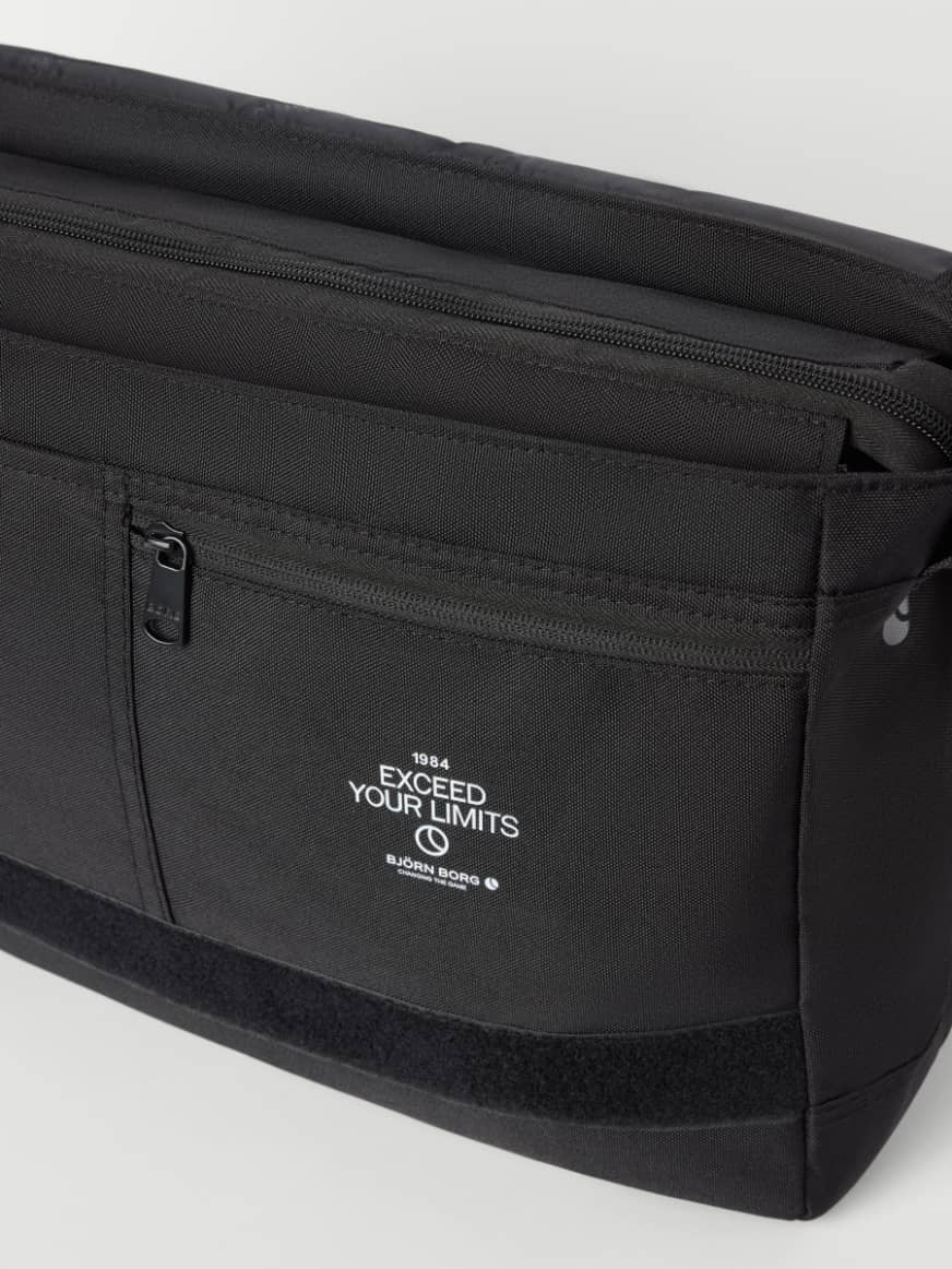 Gedetailleerde close-up van de Core Flapbag 12L in Black Beauty (Zwart) van gerecycled polyester met een rits en wit logo. - Image 3